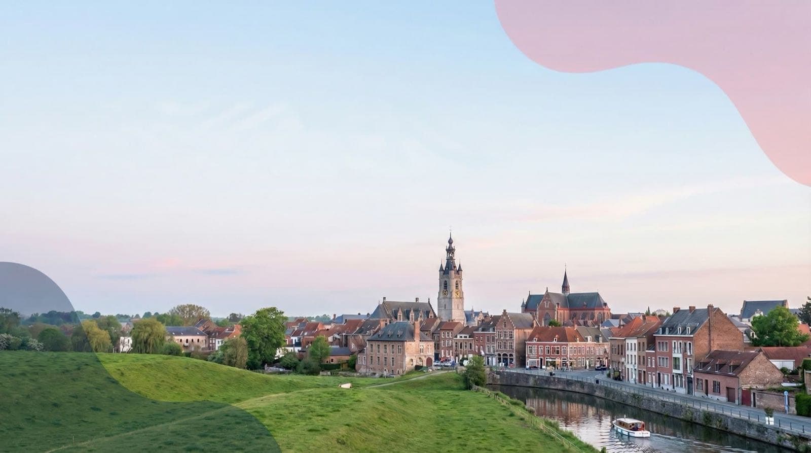 Des maisons et des bâtiments à tours bordent une rivière près d’une prairie.