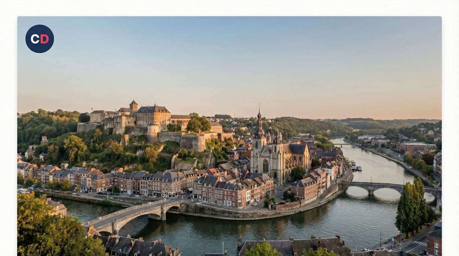 Un ensemble fortifié surplombe des maisons et un pont au bord d’une rivière.